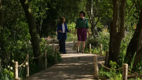 Two women are walking together along a winding, sun-dappled gravel path through a lush woodland area. The woman on the left wears a blue blazer and is gesturing while speaking, while the woman on the right is dressed in a green patterned top and purple skirt.