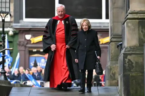 Getty Images Moira Salmond, with ginger hair and dressed in black, walks into the service with Rev Dr George Whyte, with grey hair and a red and black gown. 