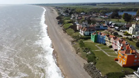 John Fairhall/BBC An aerial shot of the coast at Thorpeness, with a beach and the sea to the left and a row of houses and a colourful block of flats on the left. Beyond it is a former church, other houses. There is also a large tree-lined lake and heath land up to the horizon.