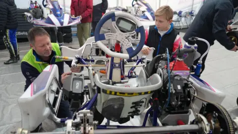 Shaun Whitmore/BBC Josh watches as his father, wearing a hi-vis top, makes adjustments to his kart. Other people can be seen working on karts behind them.