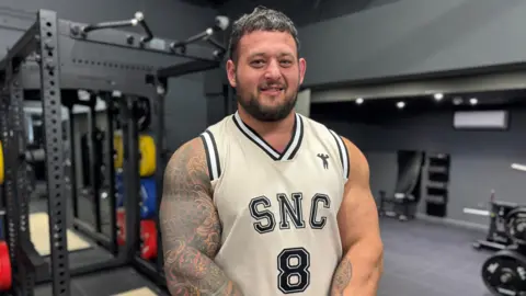 Shane Flowers stood in a gym, wearing a white vest top and sat in front of some weights