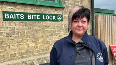 Helen has short black and white hair with hints of pink. She is wearing earrings and a blue jacket with the Conservators of the River Cam branding on the breast pocket. She is standing in front of a brick building and a fence, On the wall is a green sign reading Baits Bite Lock.