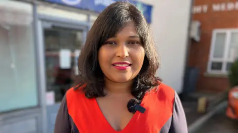 A woman with shoulder length brunette hair wearing a red and brown dress looks at the camera, while standing in front of a Boots pharmacy, with its blue signboard visible in the background.