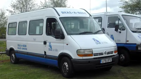 A white minibus parked on grass. The vehicle is mainly white, with a blue band around the bottom. The Dial-a-Ride logo, a hand holding a mobile phone, is on the bonnet and the door, and the phone number is on the side.