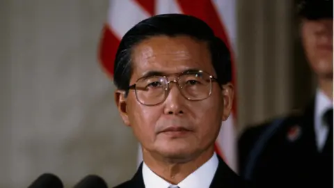 Getty Images Peruvian President Alberto Fujimori looks directly towards the camera while wearing glasses against the backdrop of a US flag.