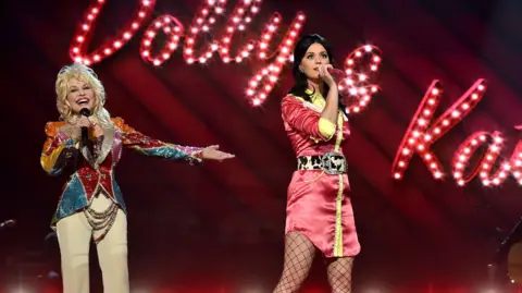 Getty Images Dolly Parton and Katy Perry singing together on stage, in front of a giant illuminated "Dolly & Katy" sign