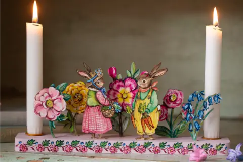 Amy Swann A long candleholder painted pink with a white candle at each end. In between is a series of brightly coloured flowers and two rabbits, one in a pink skirt and green blouse with a decorated straw hat and one in yellow overalls with a green shirt holding a bunch of carrots. 