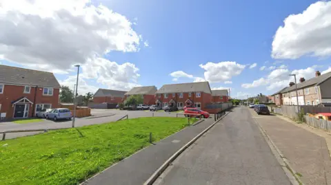 Google Raby Square looking onto Chatham Gardens. It is a set of interconnected residential streets with lined with red-bricked semi detached houses. There is a green patch of grass in the foreground.