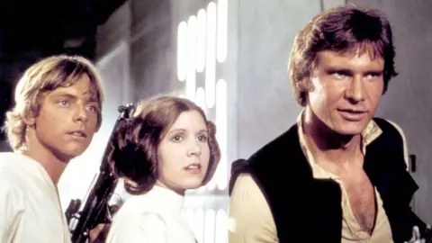 American actors Mark Hamill, Carrie Fisher and Harrison Ford on the set of Star Wars: Episode IV - A New Hope. Hamill is wearing a white top, has blond hair and is holding a black gun. Fisher is also wearing a white top and has brown hair. Ford has brown hair and is wearing an unbuttoned cream shirt with a black sleeveless top. Hamill and Fisher look concerned, while Ford looks determined.