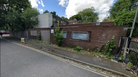 A derelict brick building can be seen with grills over windows and paint on the glass. The building is one level with an extension on the left side with two levels with one area white in colour. Trees grow over the taller building and at the front of the smaller one.
