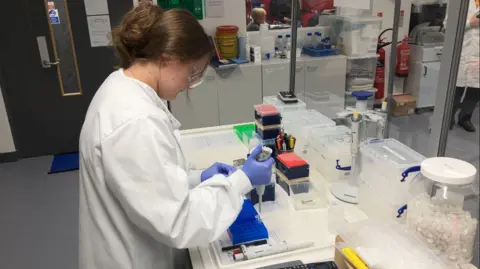 A female reseacher in a white laboratory coat, wearing blue latex gloves, putting a sample into a vial through a syringe in a clinical setting.