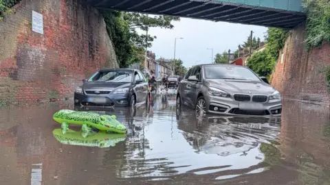 Jacob Ottaway/BBC A blue Ford car and a silver BMW car appear to be motionless in flood water below a railway bridge. In the foreground is also a green crocodile inflatable. In the background are some people and houses.