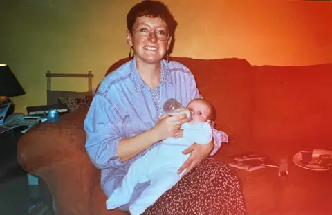 Naomi Barlow Rachel, in a denim, buttoned up shirt, feeding Naomi as a baby with a bottle of milk