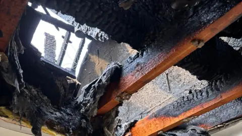 Jess Burnside Wooden beams and charred remains of ceiling insulation, and beyond that brickwork and the sky.