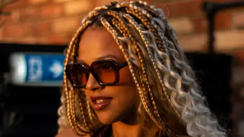 Jenna Cole Jenna Cole has braided hair which is light brown and blonde. She is wearing brown sunglasses and smiling.