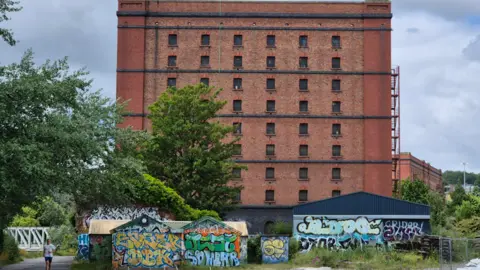 Alex Seabrook A large red brick warehouse with graffitied walls at the bottom, and a path leading up to a bridge 