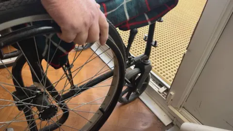 Close up of the wheels (large and small) of a wheelchair at the doorway of a house. A hand rests on top of one of the wheels. The edge of a tartan blanket is visible on the seat.