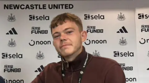 Pardoe Kendrick Pardoe Kendrick wearing a burgundy quarter-zip jumper and sitting behind a desk at Newcastle United.
