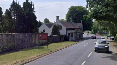 Cars drive up and down an A road in a rural area near a traffic light controlled junction on a sunny day. There is a Cotswold stone pub on the left hand side of the road with a walled garden and trees.