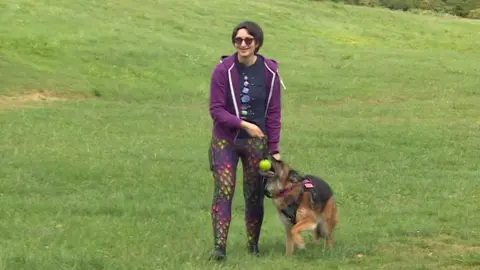 A woman in sunglasses, a purple sweater, dark tee-shirt and shiny scale-pattern leggings leads a German Shepherd dog across a park. The dog has a tennis ball in its mouth.