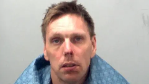 Essex Police Lee Rance has brown hair and is wearing a blue blanket around his shoulders in this picture, taken in police custody.