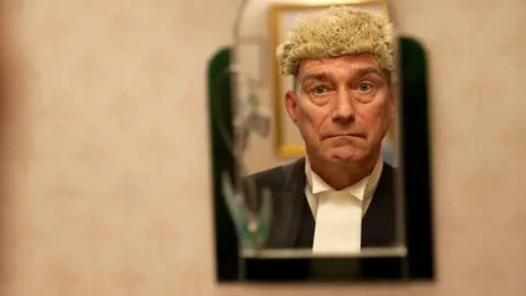 Sheriff Alastair Carmichael, a 62-year-old man, wearing a legal wig and outfit, looking into a mirror