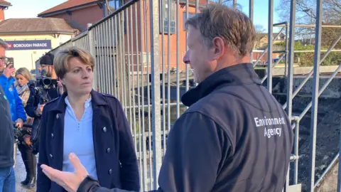 The floods minister Emma Hardy is seen standing on a bridge talking to a man wearing a jacket which reads ' Environment Agency.' There are camera crews behind her.