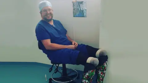 Neil Hopper is sitting in a chair wearing blue scrubs and glasses with his prosthetic limbs on show. The prosthetic limbs have a Welsh dragon pattern and are white, red and green. He is smiling at the camera. 