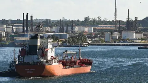 A container ship sails along choppy waters near a sprawling industrial facility close to the shore. It is daytime and a bird flies over the water.