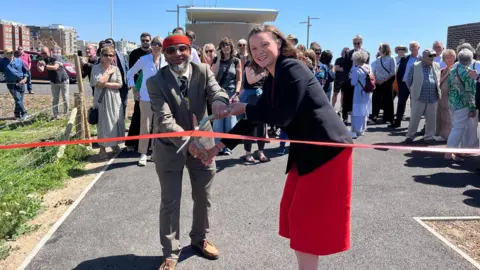 Bella Sankey cutting the red ribbon on Hove seafront with a crowd of people behind her. She is wearing a black blazer and red dress and smiling. Next to her is mayor Mohammed Asaduzzaman wearing a brown suit and red and green hat and sunglasses with his civic chain on