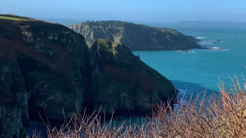 Two grass covered rocky headlands project out to the sea from the left of the shot. It is a sunny day and the sun is shining. 