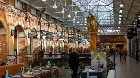 The inside of a shopping centre showing tables and archways between shops. There is a glass roof overhead and the sides of the centre are made of red brick