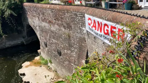 Owen Sennitt/BBC A banner stating: "Danger, keep out" can be seen attached to a bridge, with spikes placed along the top of it over a river.