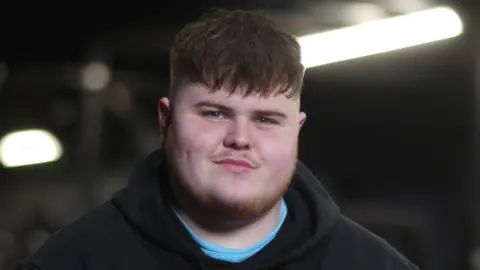 Jamie Niblock/BBC Cleverley smiles at the camera. His brown hair rests over his forehead. He wears a black hoodie with a blue top underneath.