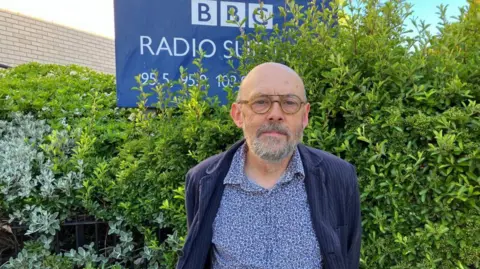 Vikki Irwin/BBC Andrew Stringer is standing in front of the BBC Radio Suffolk building. There is a hedge to the right in the background and to the left there is a BBC Radio Suffolk sign. Andrew is wearing a patterned blue shirt and dark blue striped coat.