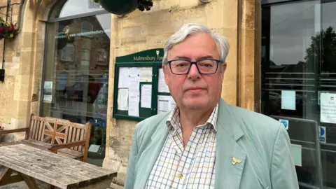 Councillor Gavin Grant stands in front of a tourist information building. A town noticeboard can be seen behind his head along with a bench with a Union Jack on it. 
