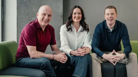 BBC Scotland Martin Geissler, Laura Maciver and Phil Goodlad sitting on green sofas. They are all casually dressed. 