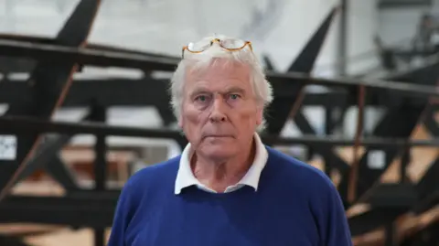 Jamie Niblock/BBC Sean McMillan, trustee and chair of SHSC, is pictured facing the camera. He is slightly smiling while standing in front of the ship replica. He has glasses resting on his head with a blue jumper and white shirt on.