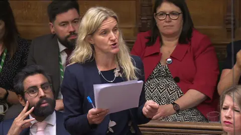 Kim Leadbeater speaking in the Commons holding some papers and a pen. She has other MPs sat around her.