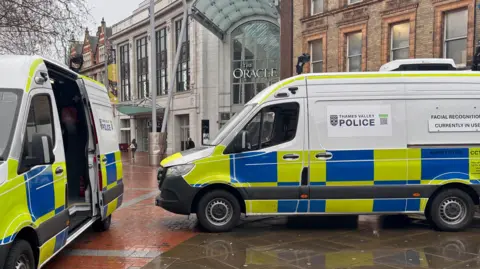Two police vans, each with two facial recognition cameras mounted on the roof are parked in the pedestrianised Broad Street outside the Oracle shopping centre entrance in Reading. The sky is grey and the pavement is wet.