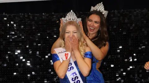 Claire Redgrave and Lauren Jennings pictured in front of a sparkly black background. Both are wearing sequined blue dresses. A smiling Lauren is putting a tiara on Claire's head, while Claire has her hands over her mouth in disbelief. Claire has long blonde hair and has a Miss Essex sash on, while Lauren has long brunette hair.