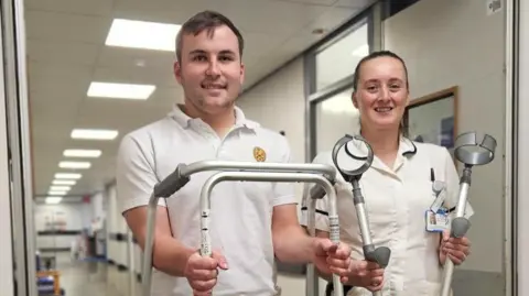 A man and women in a hospital corridor, he in a white T-shirt, she in a white hospital shirt. He has short brown hair and is holding up a walking fame. She has brown hair tied back and holds two elbow crutches. Both are smiling.