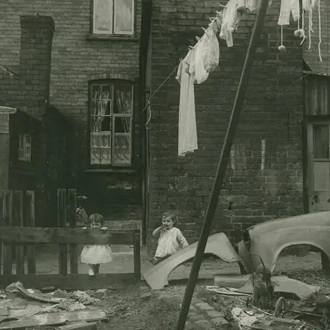 Estate of John Blakemore provided by Coventry University An image of two young girls stood outside an old terraced house with a washing line hanging a number of white items of clothing