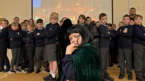 BBC A group of primary school aged pupils pose for the camera with one boy dressed as Claudia Winkleman in a wig and cloak