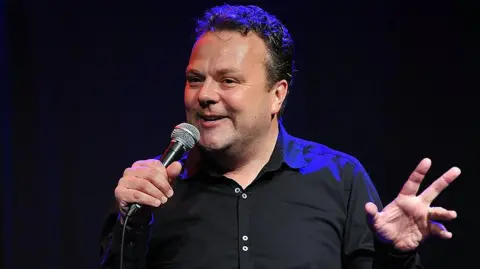 Getty Images Hal Cruttenden in black shirt looking out to a crowd holding a microphone.