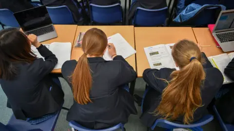 PA Media Tiga gadis sekolah menengah duduk di meja di ruang kelas. Mereka memiliki punggung ke kamera. Mereka menulis di makalah tes.