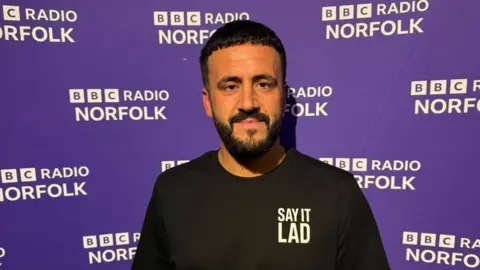Rob Butler/BBC Danny Tanti has short black hair combed forward, and a black beard and moustache. He is wearing a Say It Lad branded tee shirt, and is standing in front of a BBC Radio Norfolk branded wall.