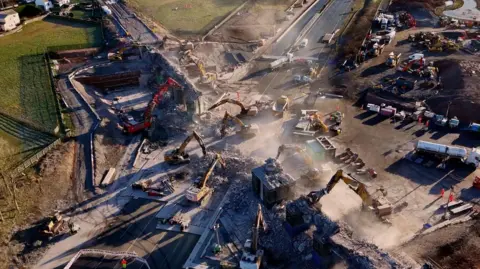 A seven diggers are demolishing a bridge over the M6. A dust cloud is hanging over the construction sire and construction vehicles are waiting to carry rubble away.