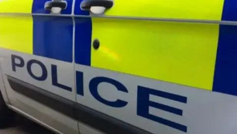 The side of a police car, with POLICE written across the side and the usual blue and yellow markings