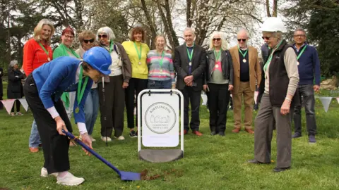A small group of smiling people watch as a woman, wearing a blue hard hat, begins to dig a patch of grass, with a blue spade. They are all stood on a grass filed with trees and colourful bunting behind them. Only a tiny patch of ground has been disturbed by the digging. There is also an white shop sign on the grass which reads Kirtlington Community Shop & Cafe